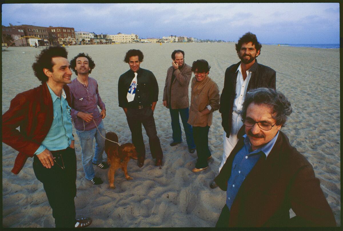 Seven male architects, some in blazers, and one holding a dog on a leash, stand on a beach on a windy day.