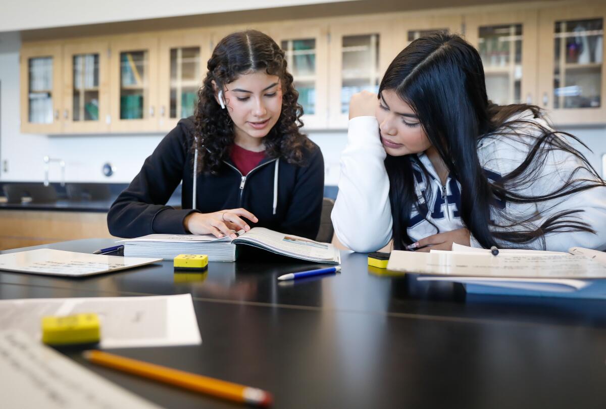 Two students sit behind a dark-color desk and look at the same book. Books, papers and a pencil lay on the desk. 