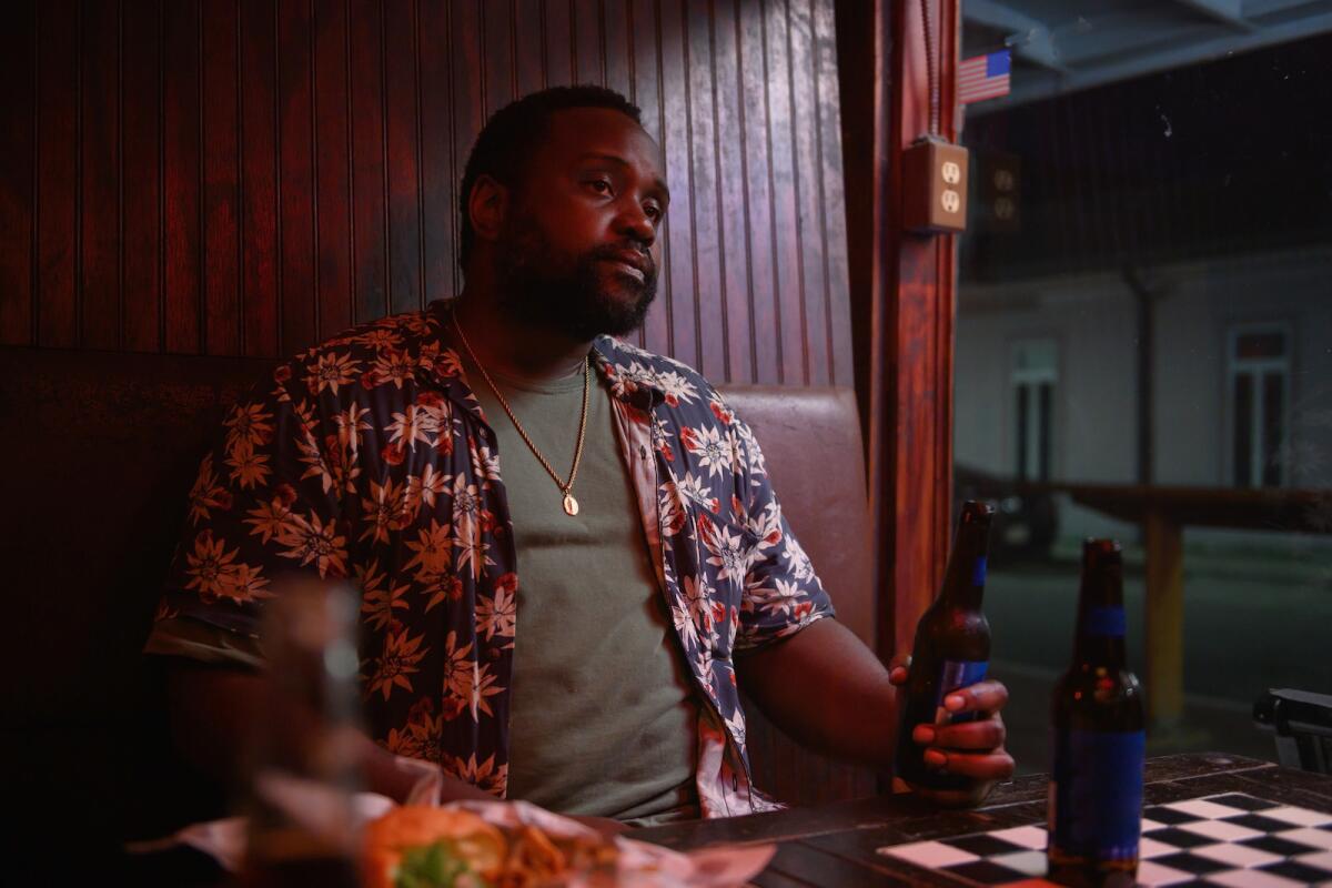 A man sits at a table, holding a bottle, in the movie "Causeway."