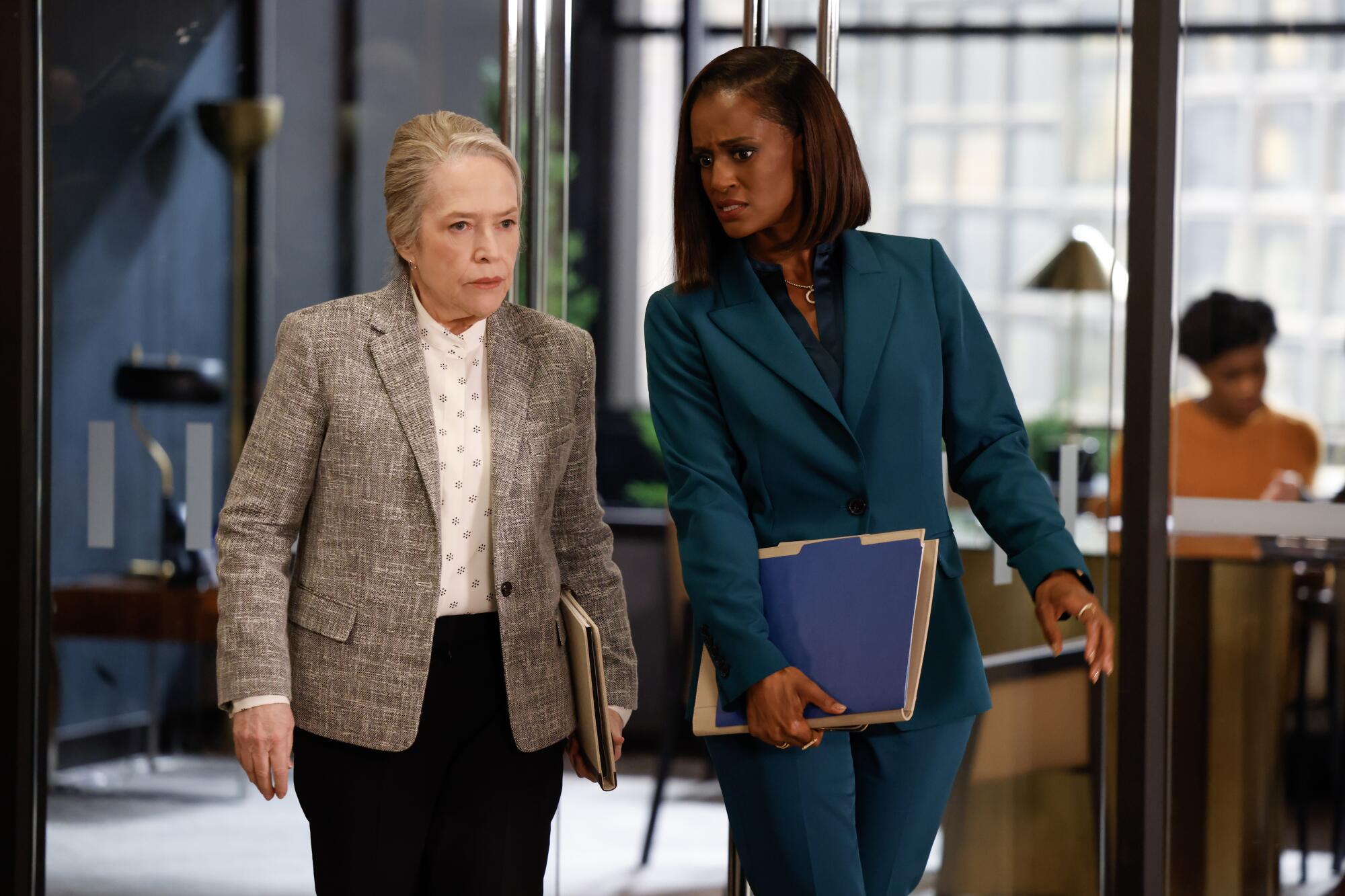 A woman in a blazer and dark pants walks next to a woman in a blue suit holding files in an arm.