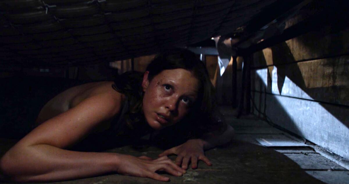 A young woman huddles under a bed with an anxious look on her face.