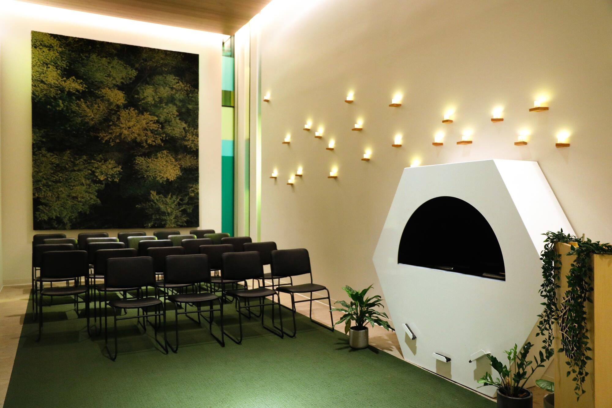 A white hexagonal pass-through vessel sits along a wall near folding chairs lined on a green carpet.