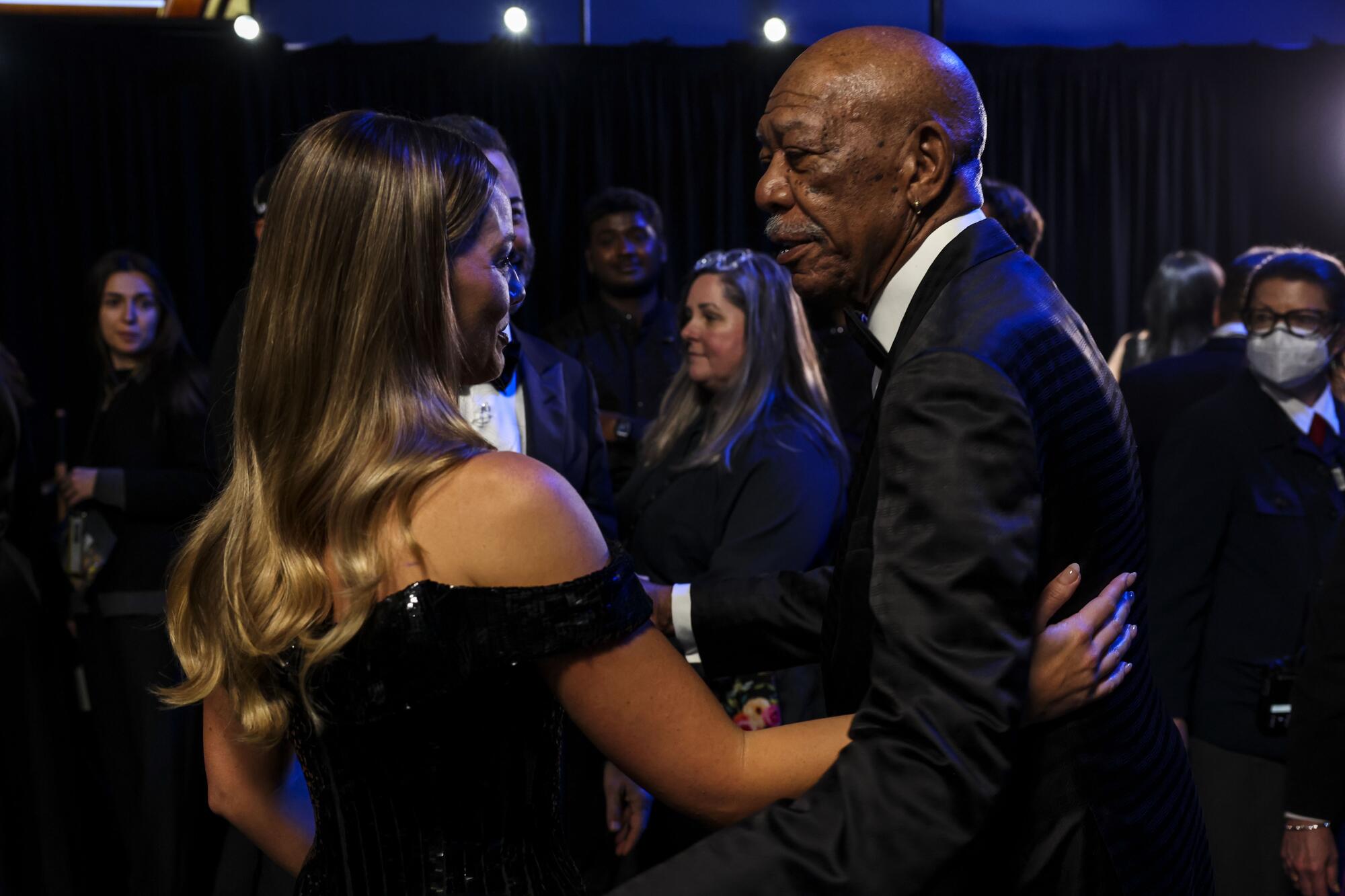 A woman in a black dress chats with a man in a tux backstage at the Oscars. 