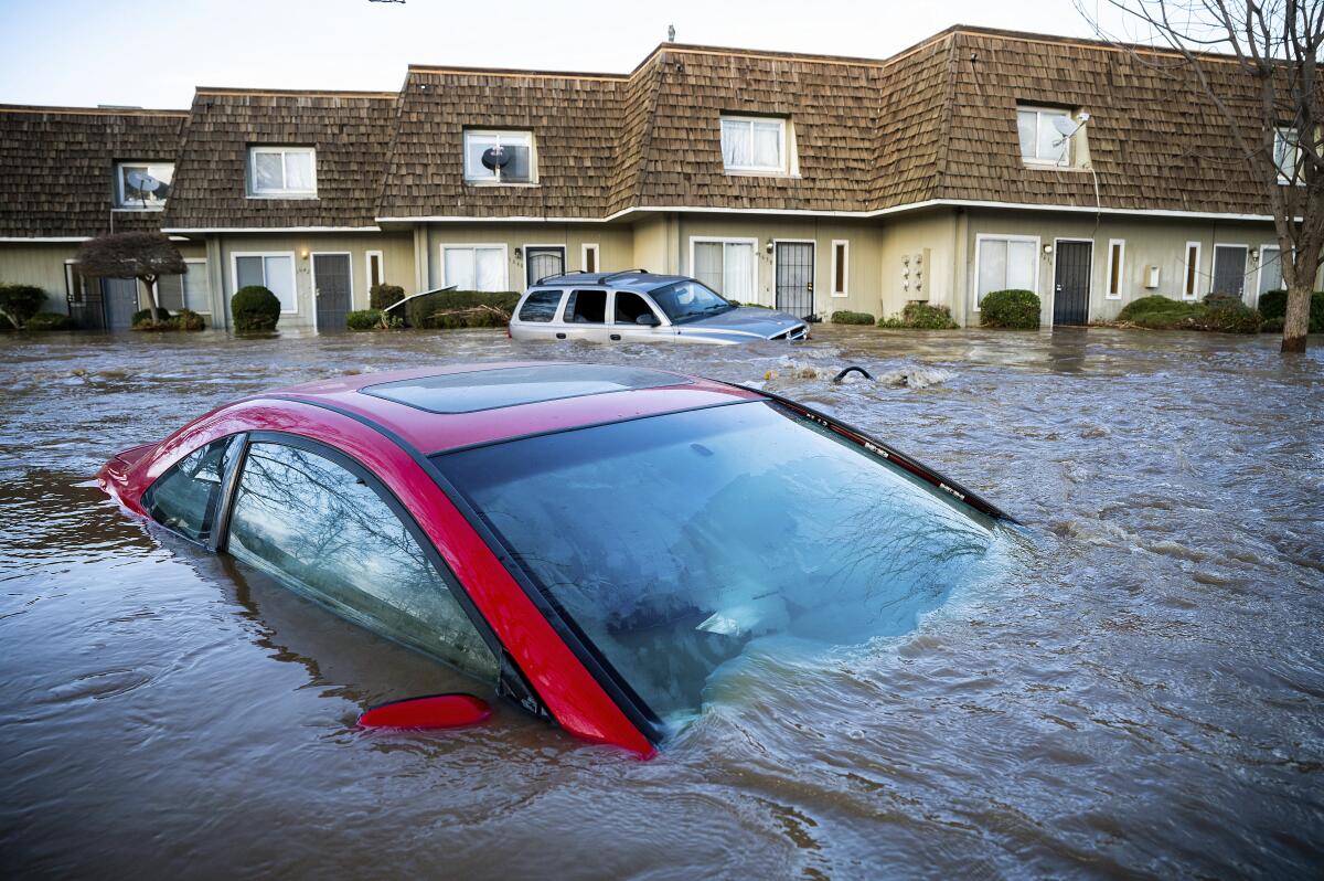 A car is submerged in floodwater up to its windows. Another flooded car and a row of townhouses are in the background. 