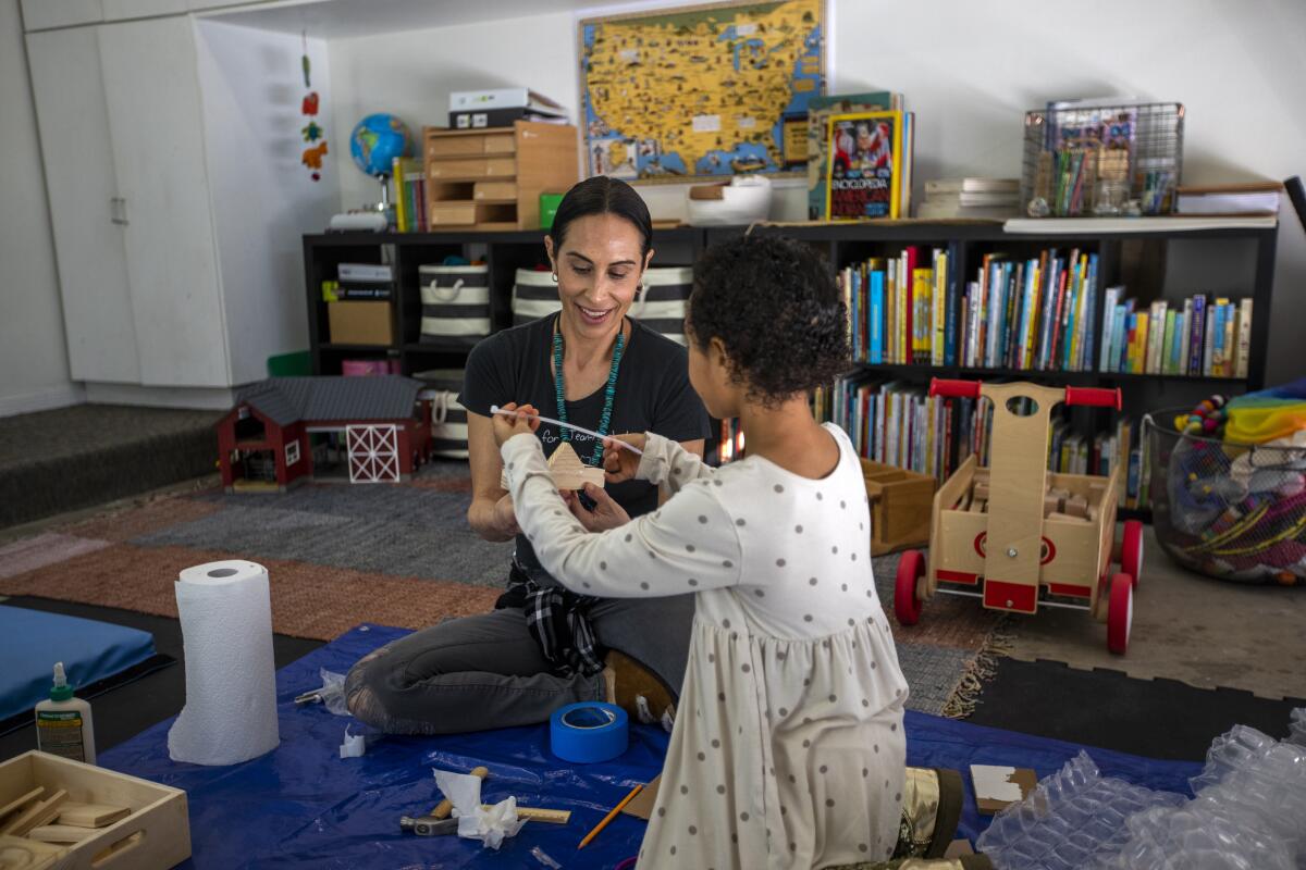 A woman sits on the floor of a room with books and maps, building something with wood and glue.