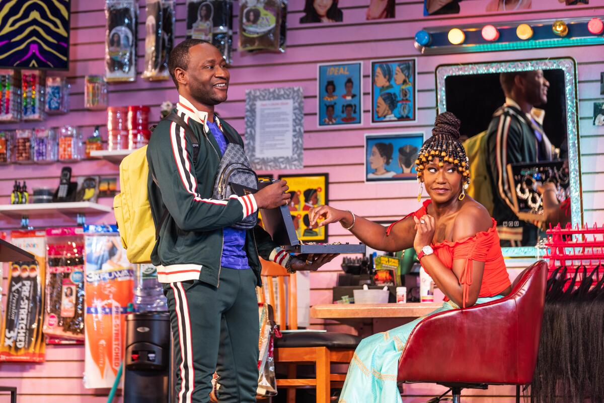 A standing man in a track suit and a seated woman with elaborately braided hair on a stage set 