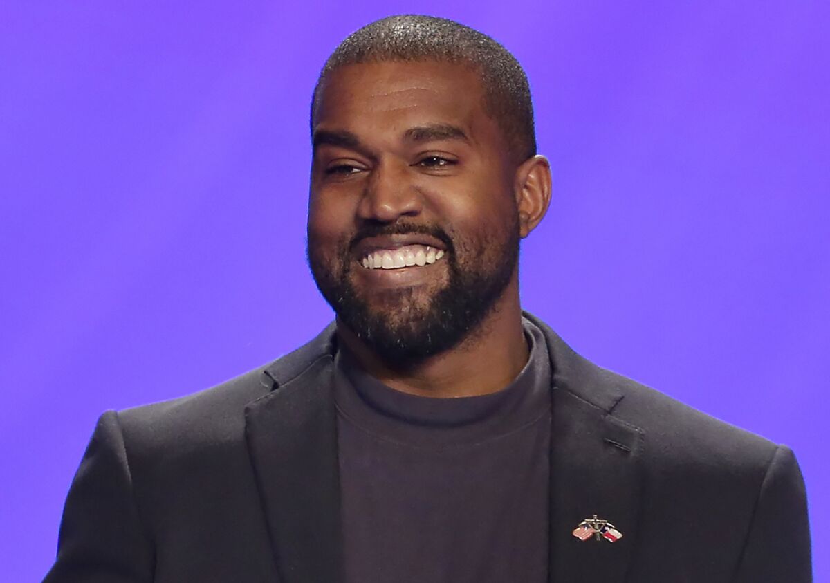 Kanye West smiles wearing a black shirt and black suit against a purple background