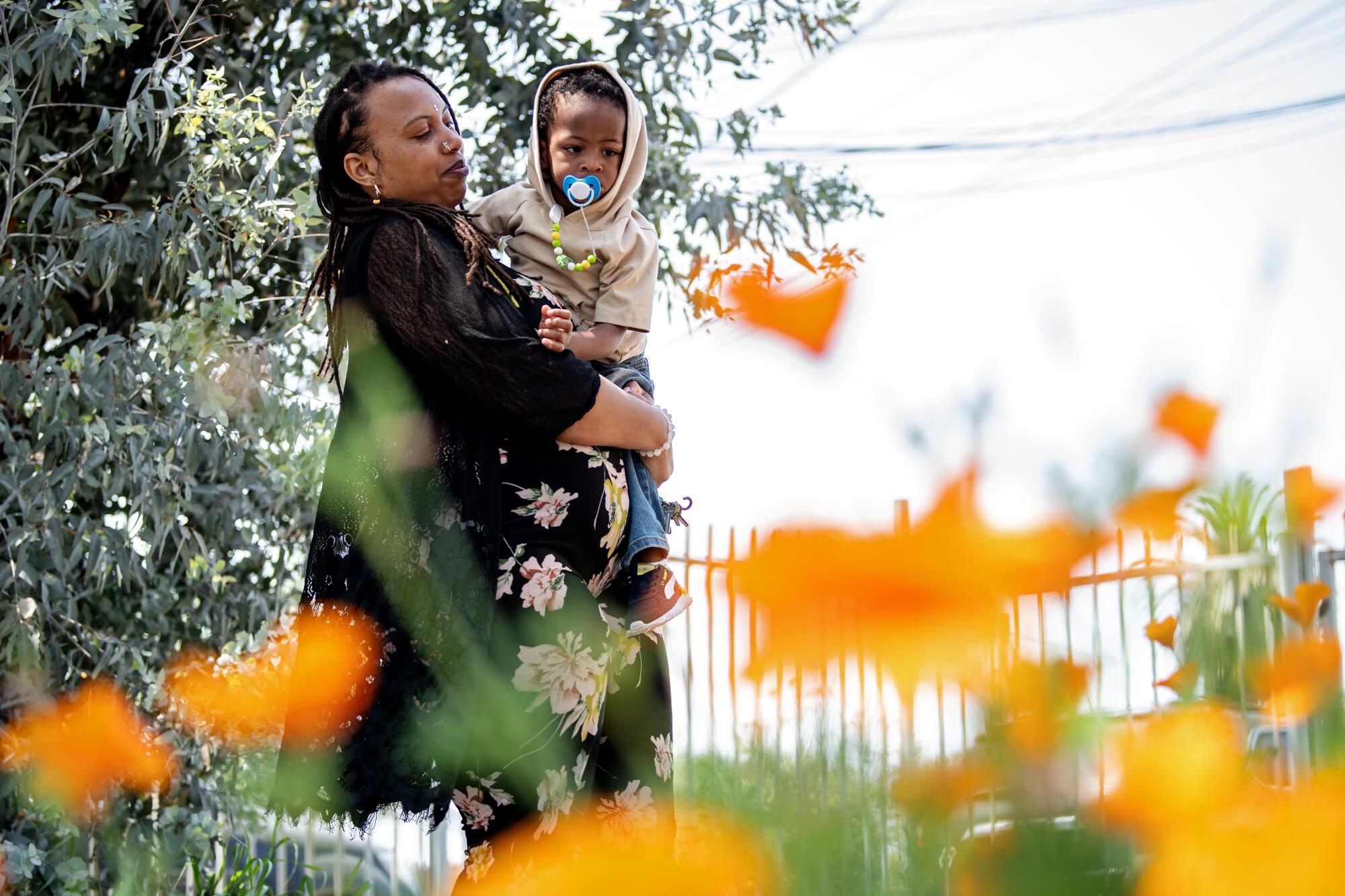 Oya Sherrills carries her 2-year-old son in front of orange flowers.