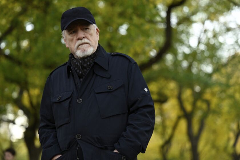 Brian Cox (as Logan Roy) frowns while wearing a black cap and jacket in the woods.
