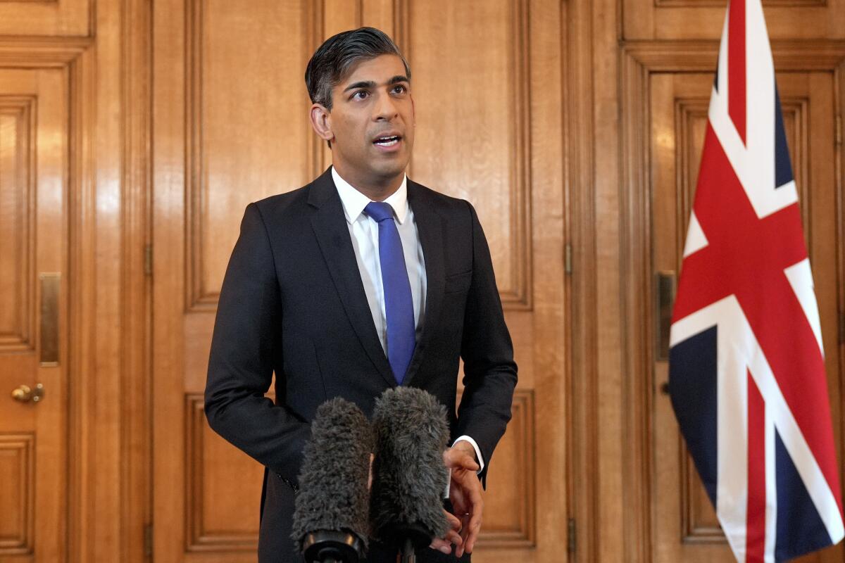 British Prime Minister Rishi Sunak stands and speaks in a wood paneled room. 
