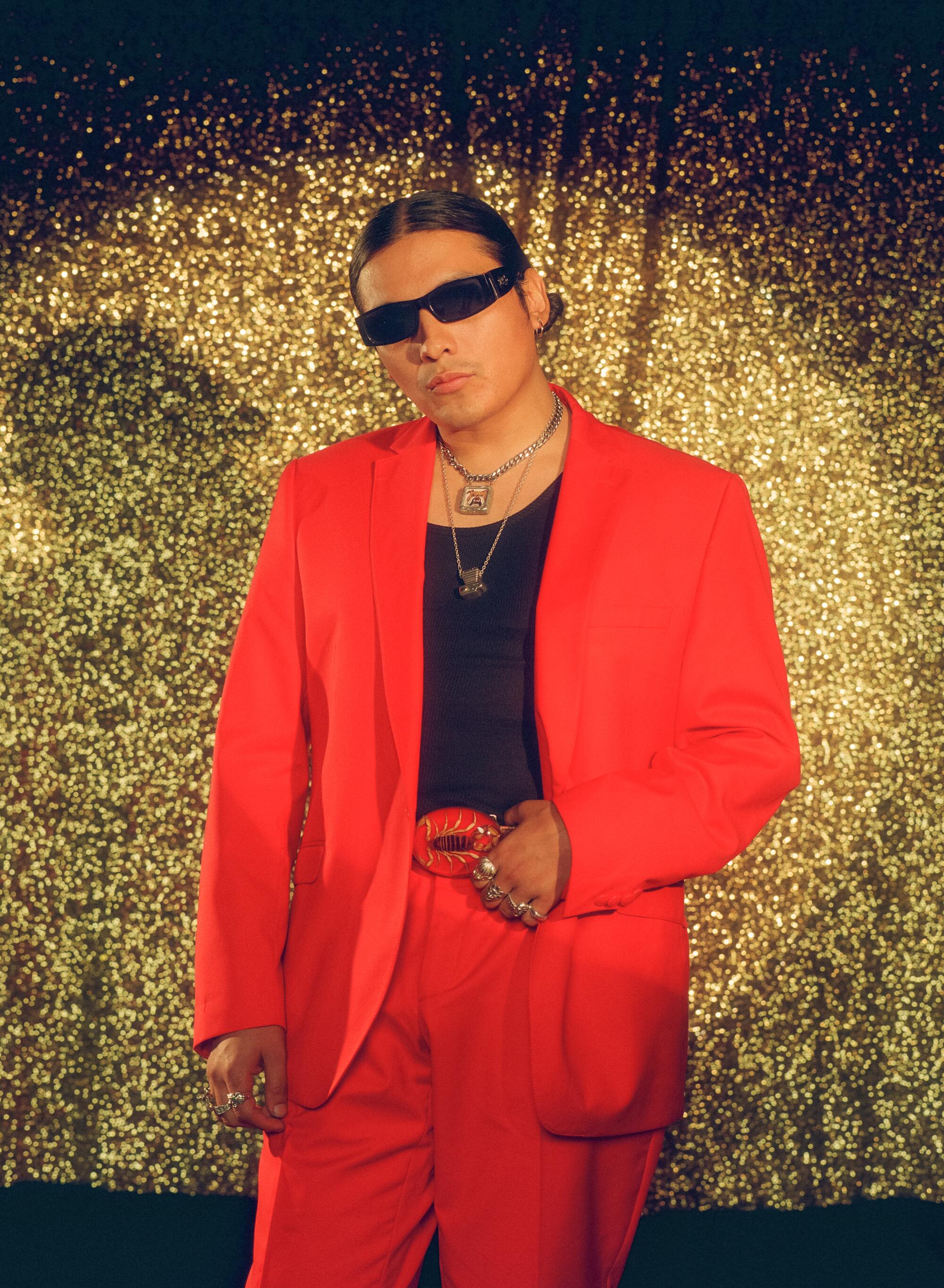 A man in a red suit and sunglasses poses in front of a golden curtain