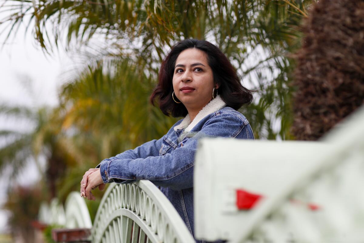 A woman with hoop earrings and a denim jacket standing at three-quarter profile with her arms on a gate