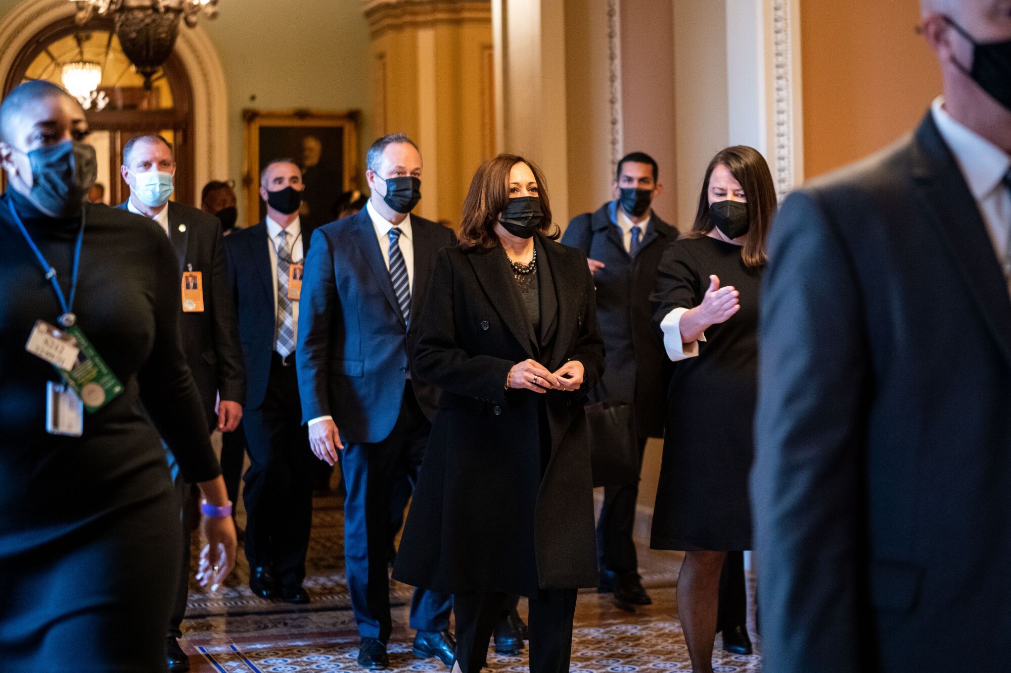 A group of men and women in dark suits and face masks walks down a hallway.