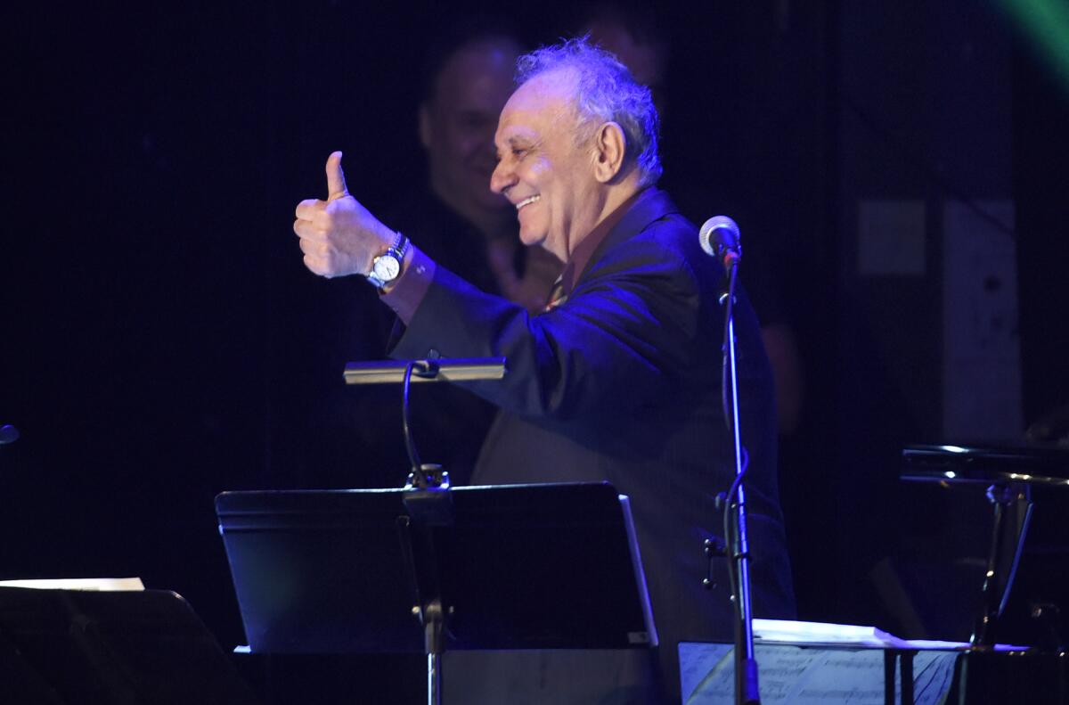 An older, smiling man looks to the side and gives the thumbs-up sign during a musical performance