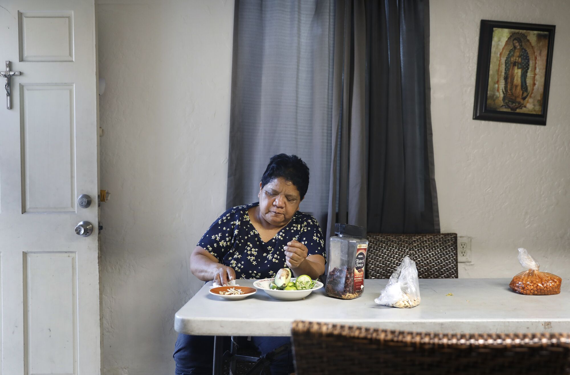 A woman sits at a kitchen table with plates of food in front of her. A picture of the Virgin of Guadalupe hangs on the wall. 