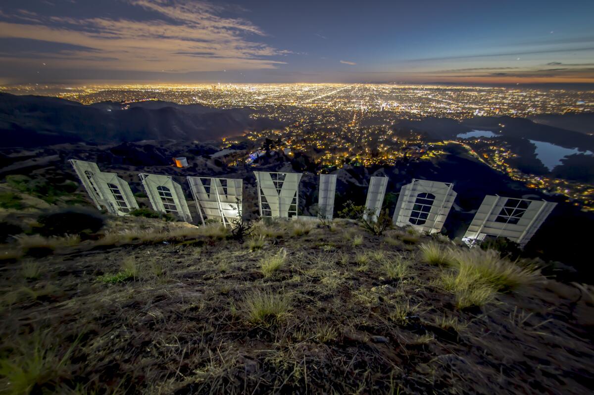 A photo taken from behind the Hollywood sign at night, with the lights of the city of Los Angeles below.