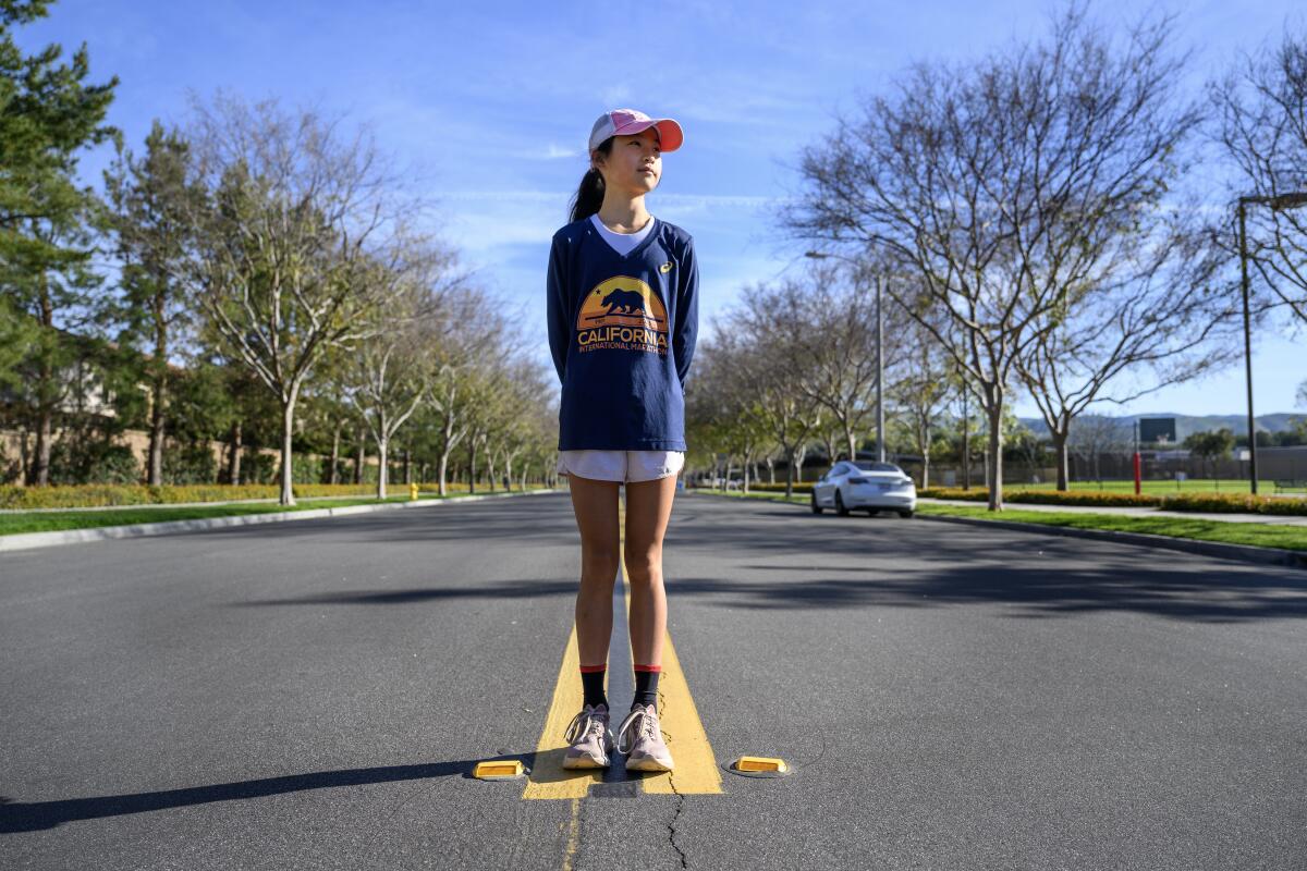  A girl in a cap and a blue long-sleeved shirt with yellow design stands in the middle of a tree-lined street