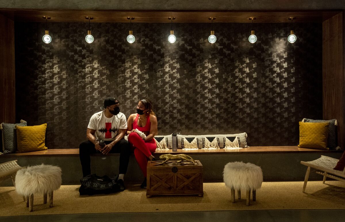 A man and a woman sit on a long bench with throw pillows and shag ottomans
