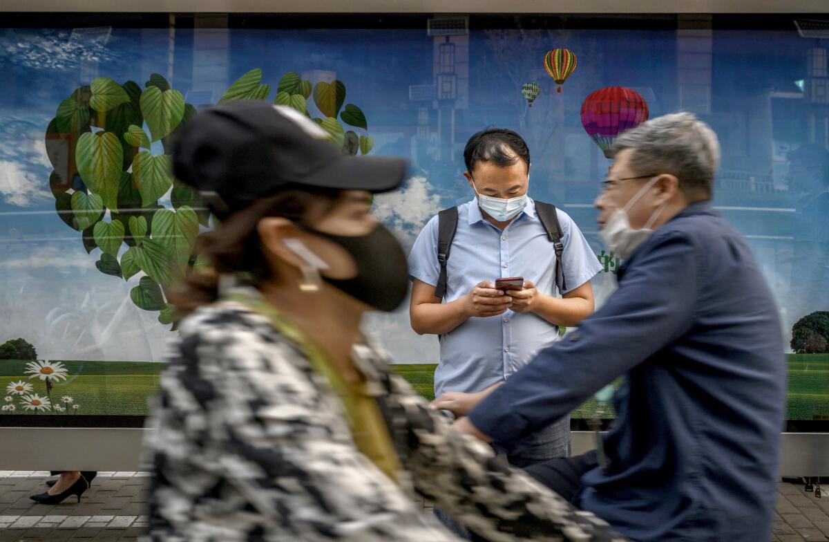 A masked man looks down at his cellphone as a woman and a man cross paths in front of him
