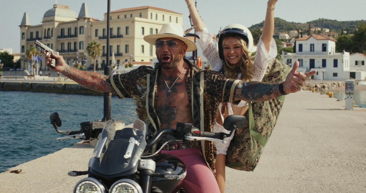 A man in a hat holding a gun and a woman in a helmet riding a motorcycle next to a body of water.