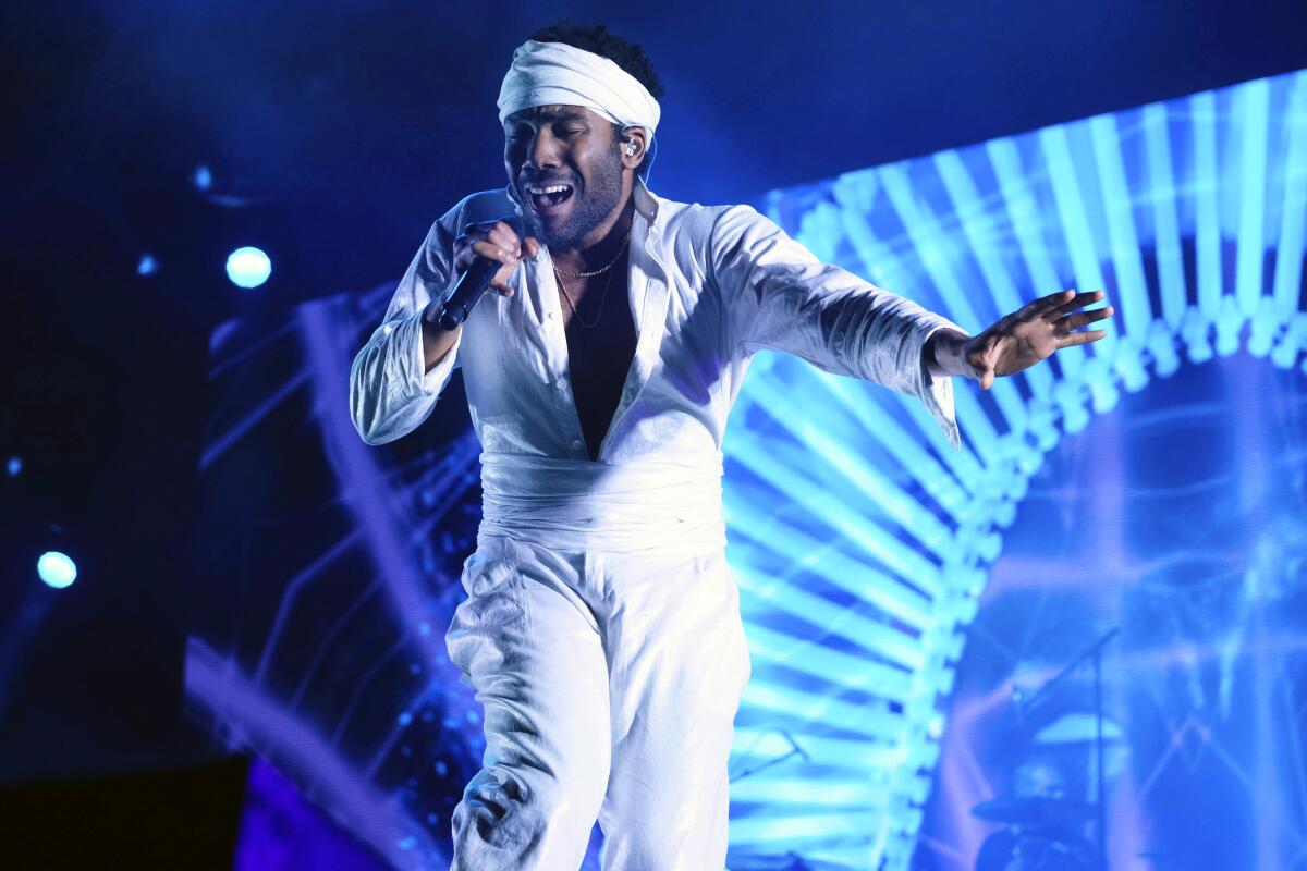 Donald Glover singing onstage in white pants, shirt and and a head wrap with blue lights behind him