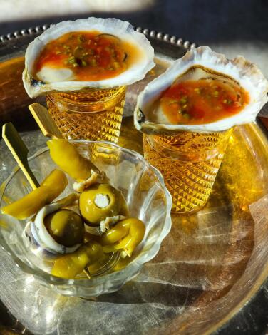 Oyster shooters on a silver plate with a glass dish that holds pickles and olives