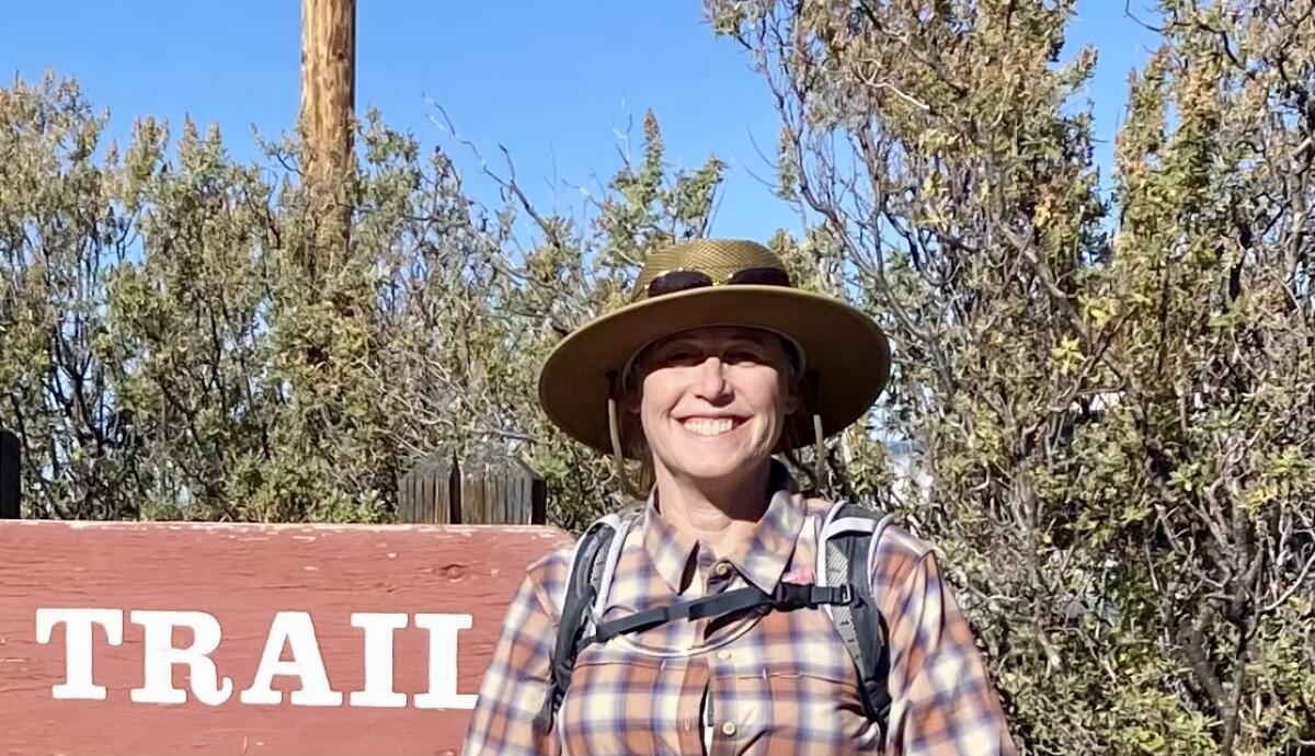 A woman in a flat brimmed hat and backpack stands near a wooden sign that says "Trail."
