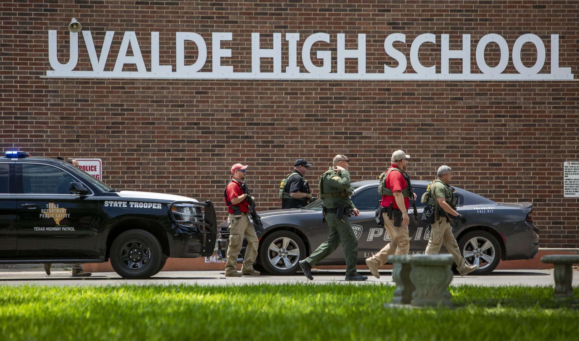 Five men walk near law enforcement vehicles parked in front of a wall bearing the words Uvalde High School 
