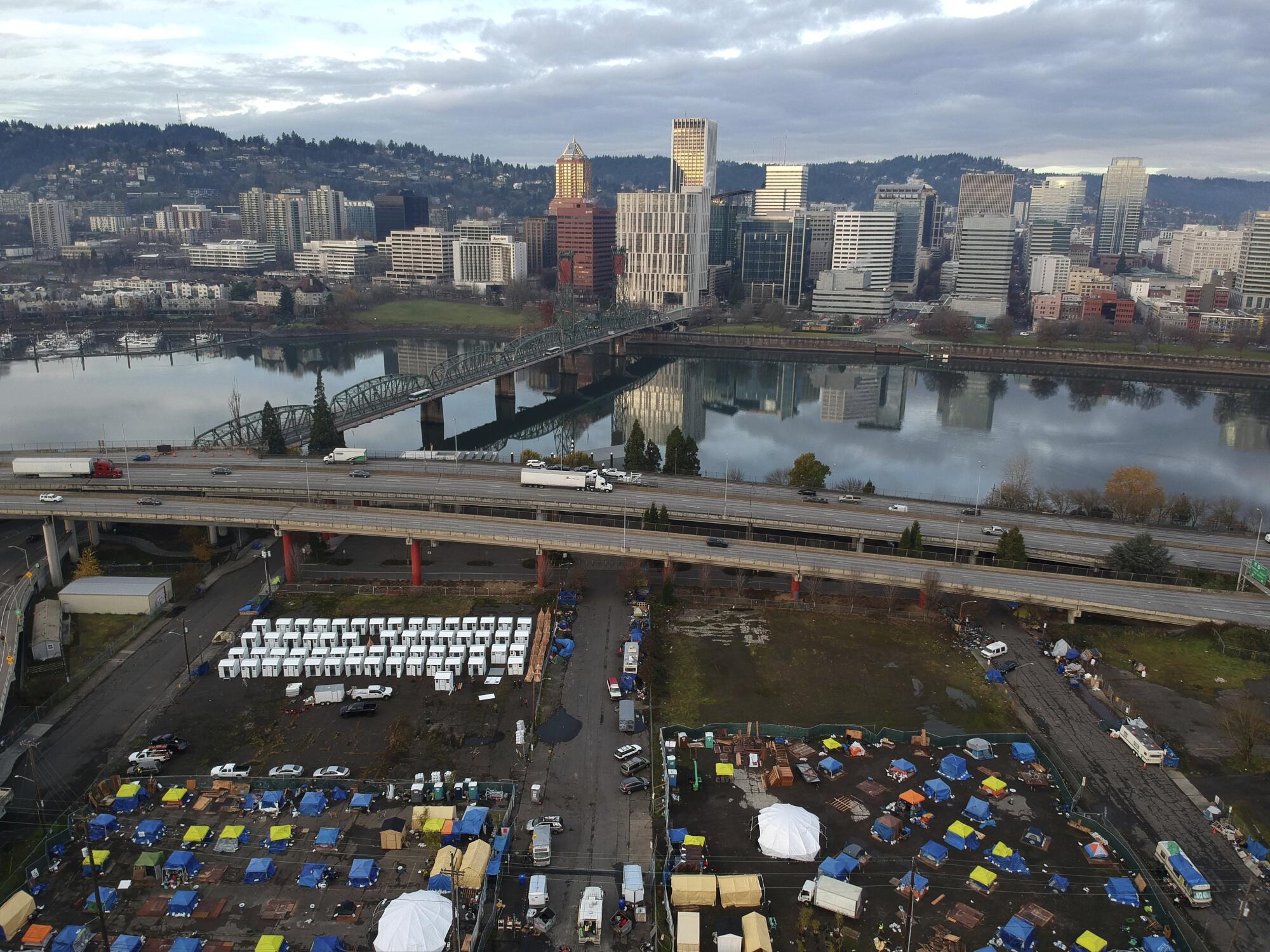 An aerial photo shows tents filling an empty Portland parking lot next to an elevated road alongside a river.