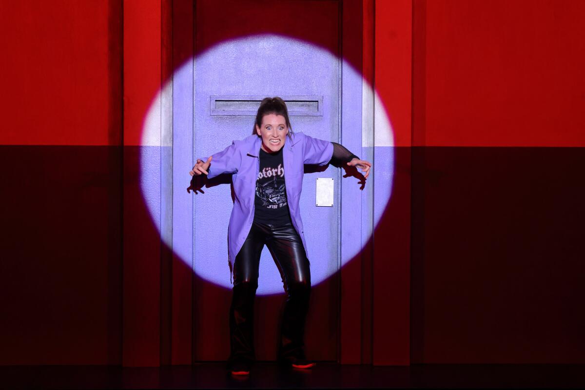 Naomi Louisa O’Connell in a purple blazer in front of a purple jail cell door in the spotlight.