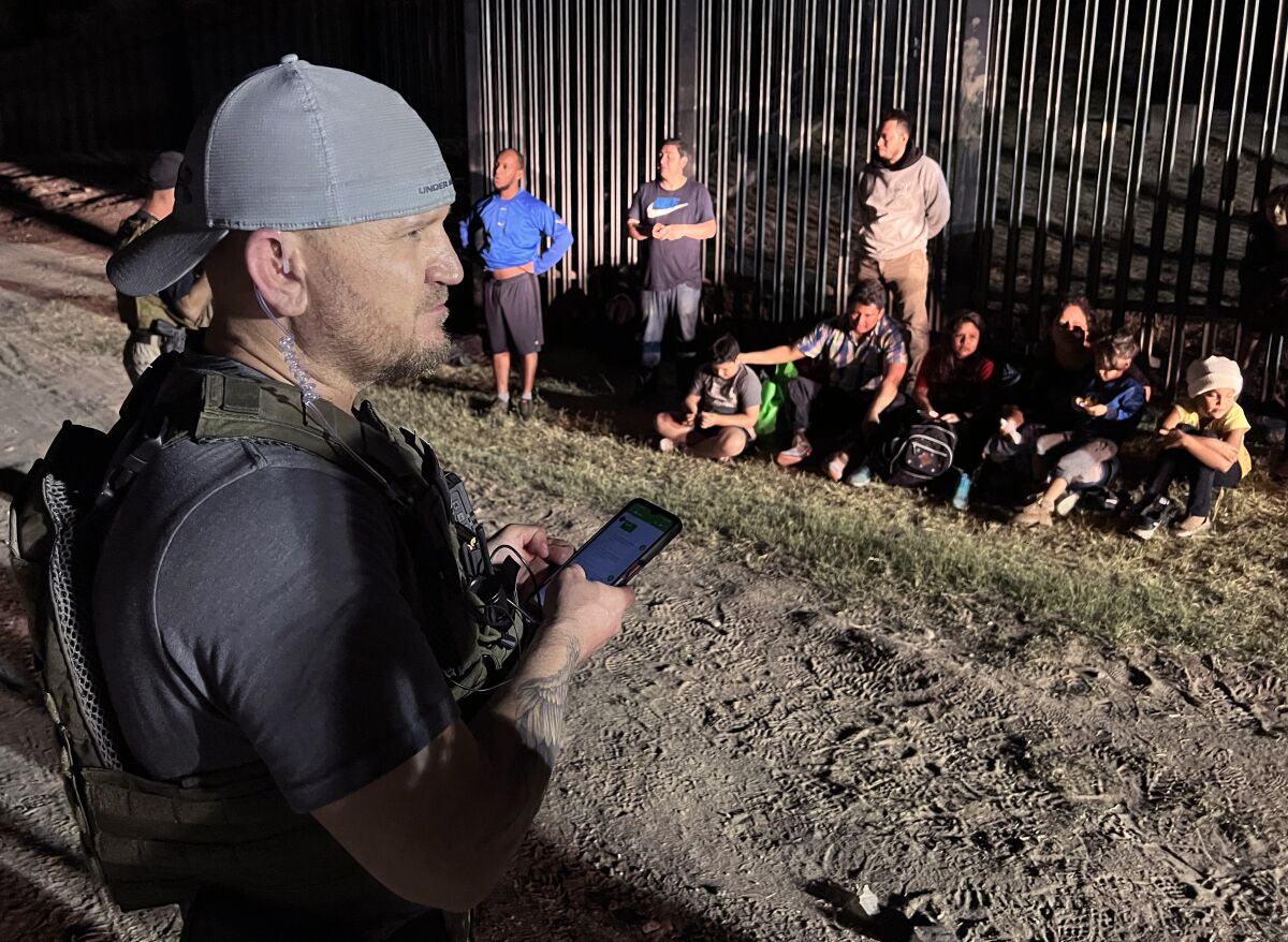 A man in militia gear standing by as several adults and children wait next to a wall