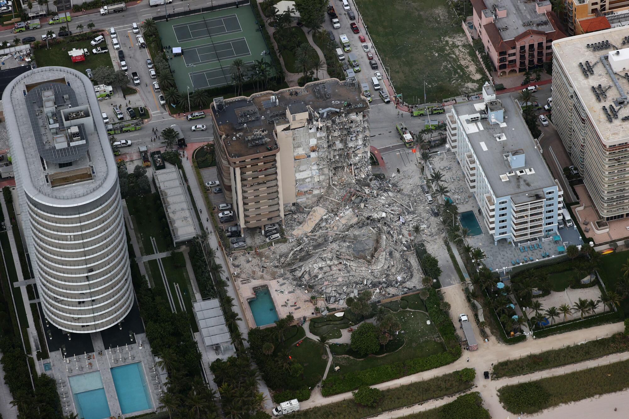 An aerial view of rubble amid multistory buildings with swimming pools and green lawns.