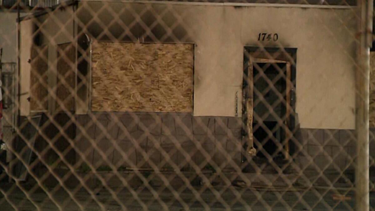 An exterior of a boarded-up building; framed through a fence and photographed at night. 