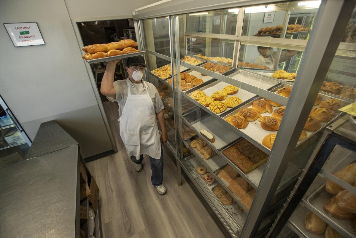 A man in an apron holding a tray of rolls overhead walks toward a bakery display case.
