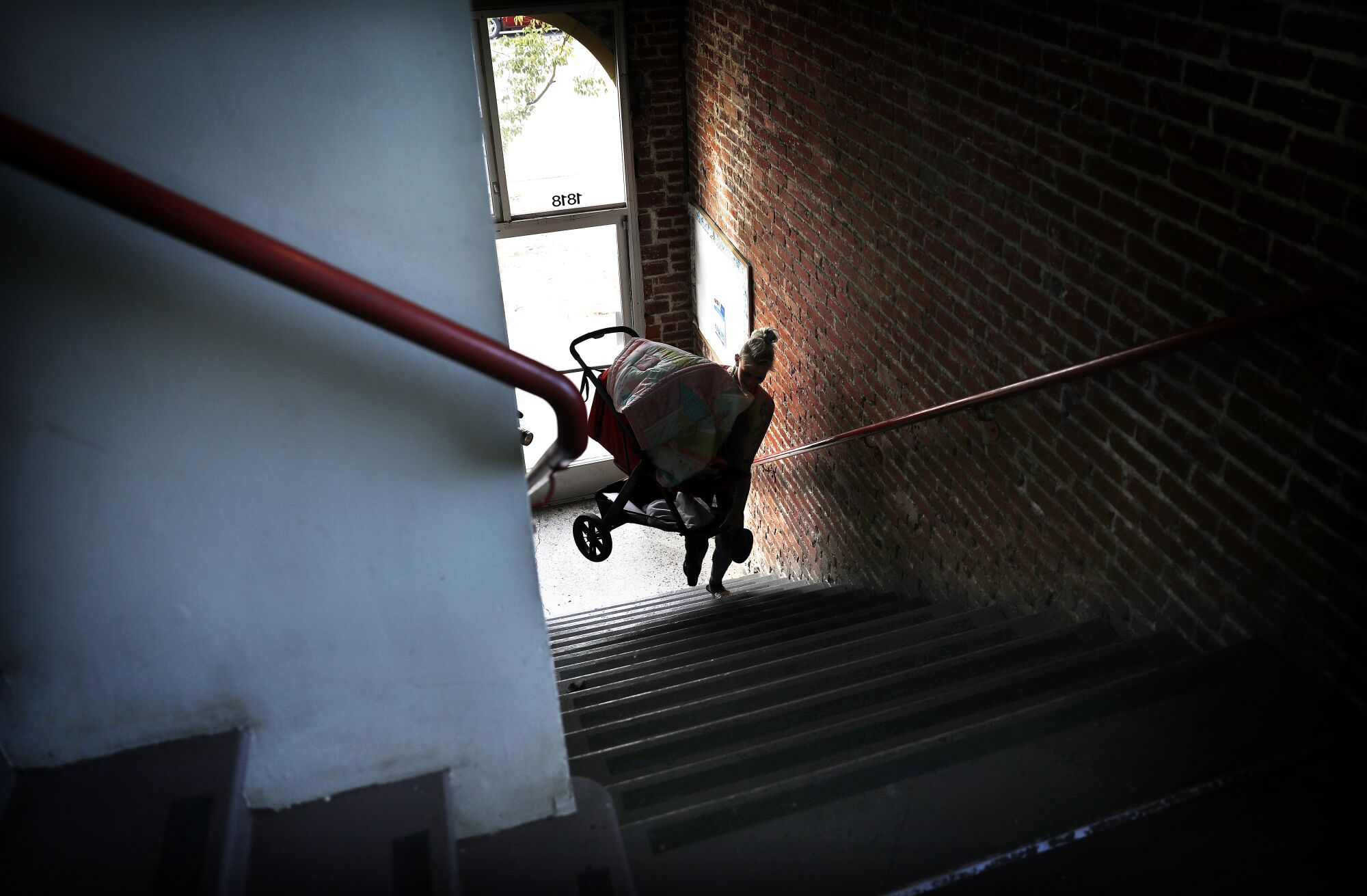 A woman backlit by a window climbs a flight of stairs carrying a baby in a stroller.