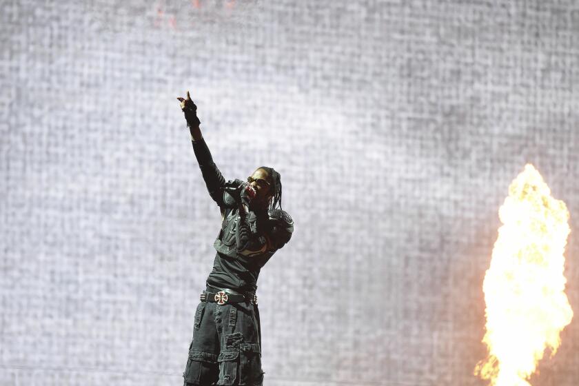 A distant shot of Travis Scott on a stage holding up his right hand and holding a microphone in front of a flame