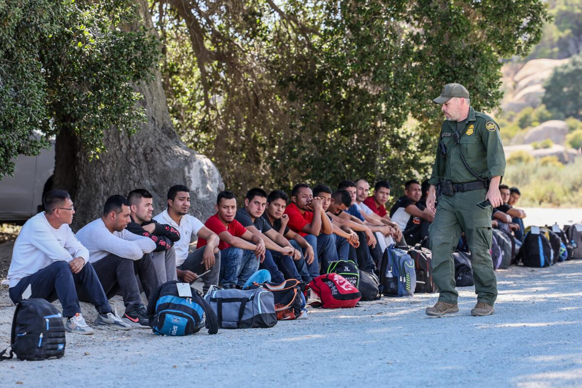 A row of men seeking asylum sit by a road next to an officer.