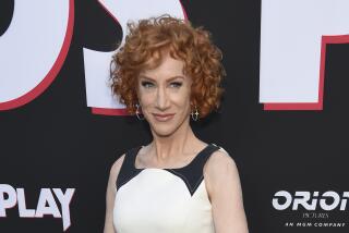 Kathy Griffin smiling with short curled red hair and wearing white and black dress, hoop earings at red carpet event