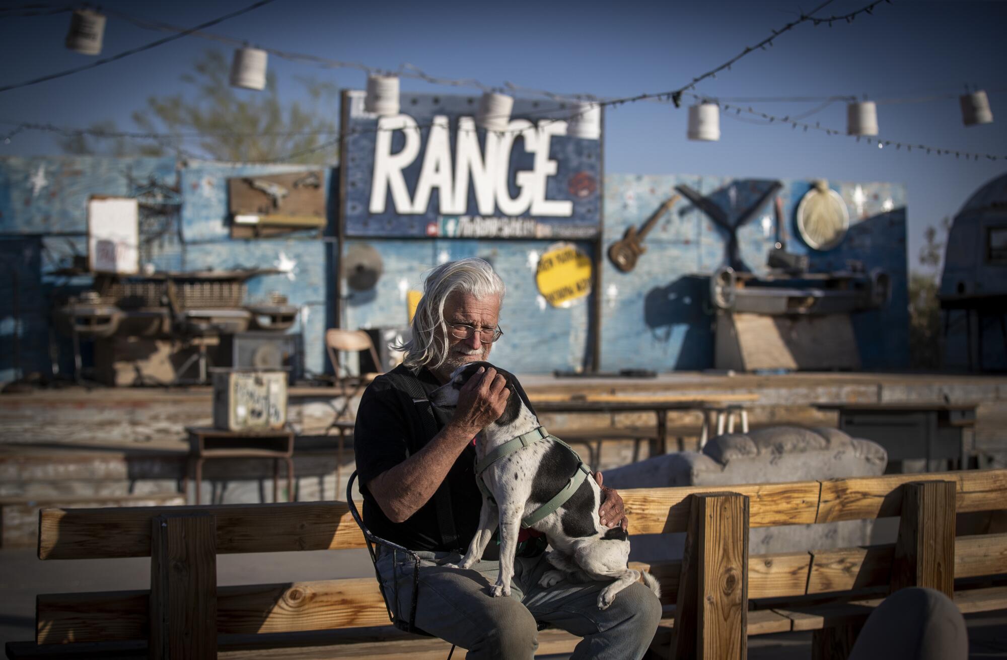A man sits on a chair with a dog in has lap at an outdoor music stage with a sign that says "the Range"