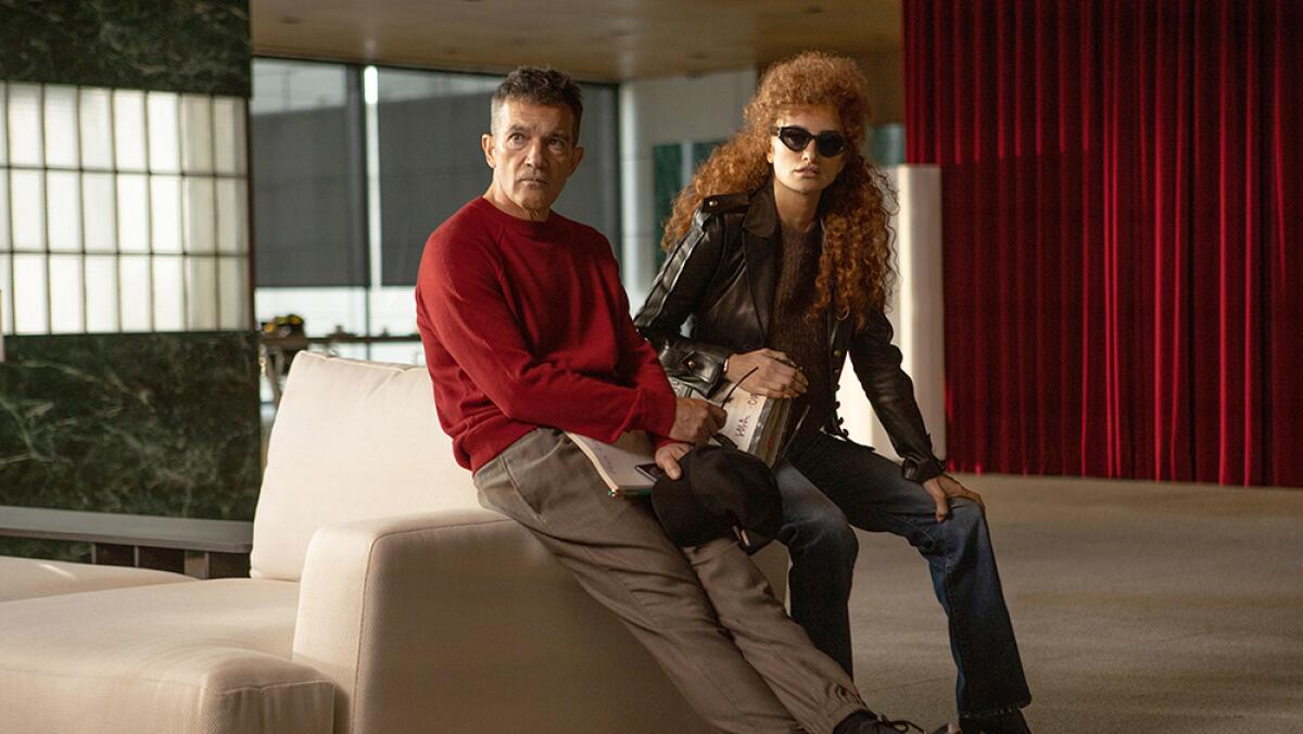 A man in a red sweatshirt leans against a white couch alongside a woman in sunglasses and leather jacket.