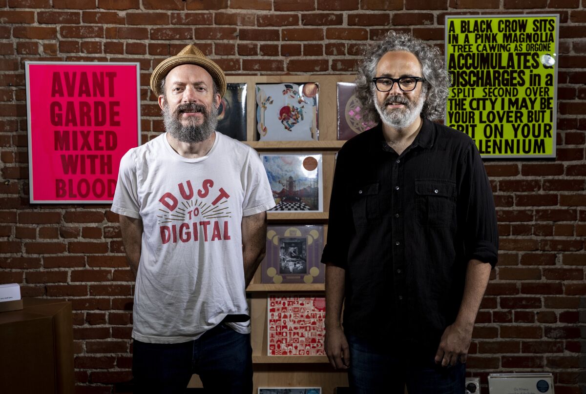 Two men stand before a brick wall with neon-colored signage and an array of records
