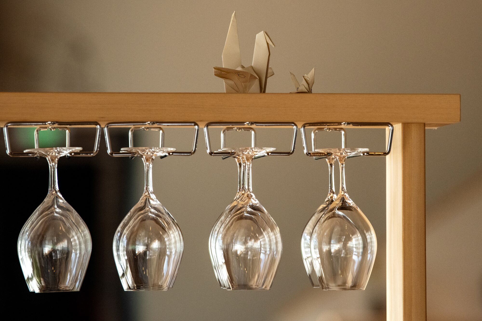 A pair of paper origami cranes atop a wooden shelf with wine glasses hanging from it.
