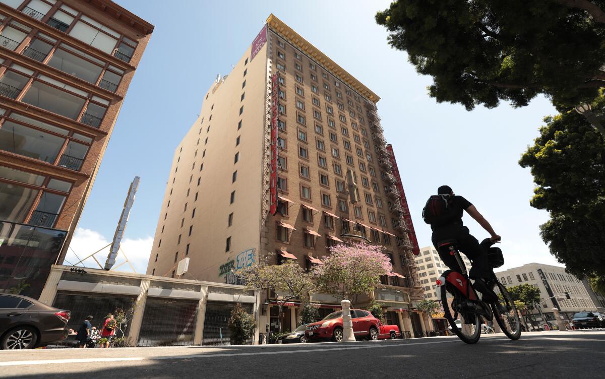 A older 15-story tan building with red signs seen at an angle from a city street, with a cyclist riding in the foreground
