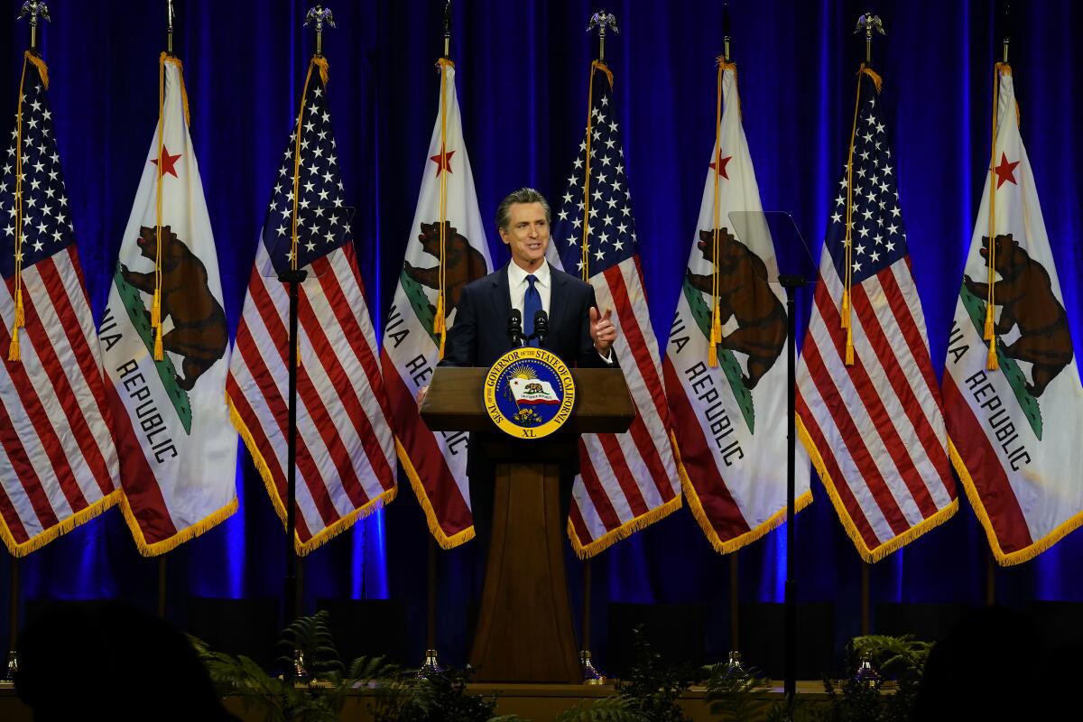 A man stands at a lectern in front of a row of American and California flags.