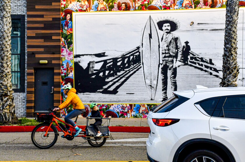 A bicyclist pedals past a mural featuring Emiliano Zapata standing on a pier holding a surfboard.