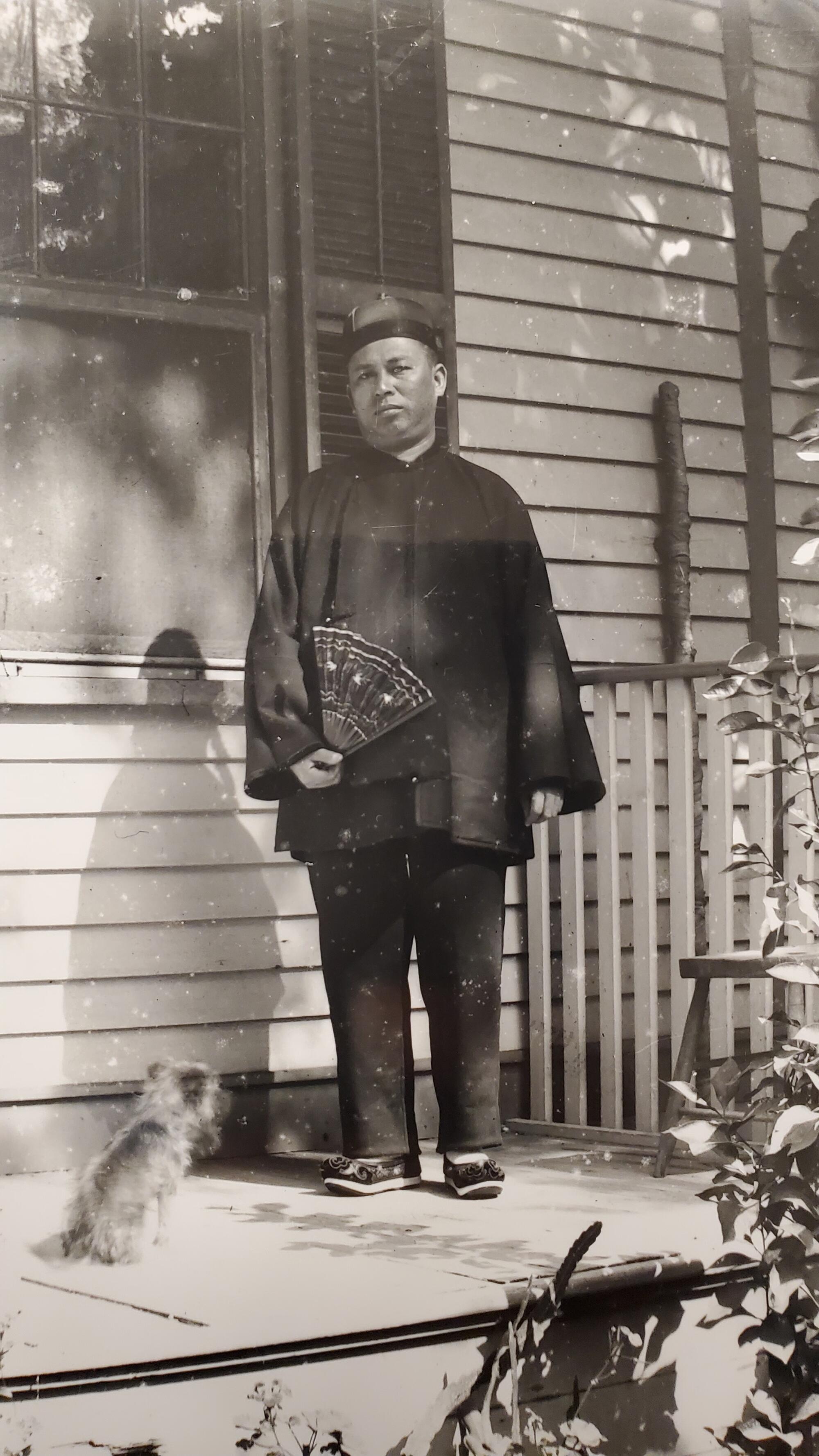An old black-and-white photo of a man standing on a porch, a small dog at his feet.