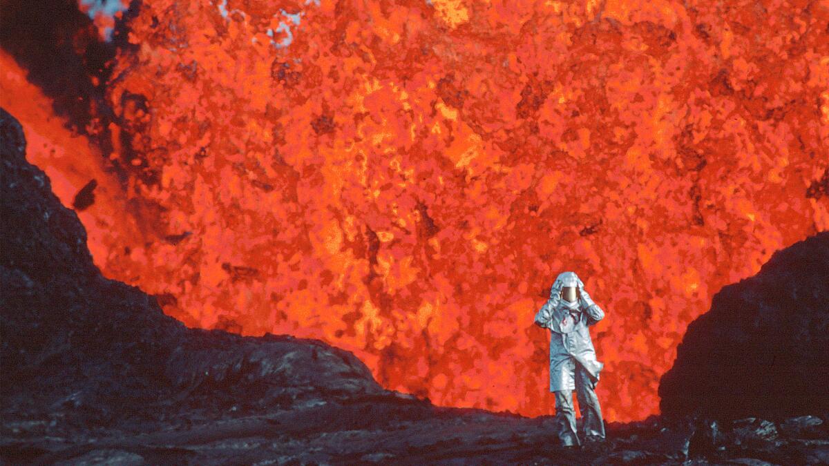 A figure in safety gear stands close to lava shooting up from a volcano. 