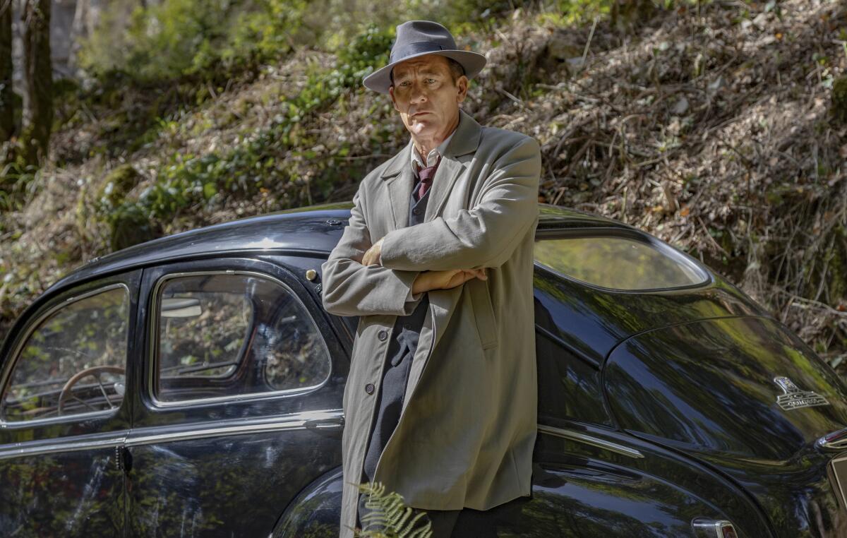 A man in a grey trench coat and hat leans against a black 1940s car.