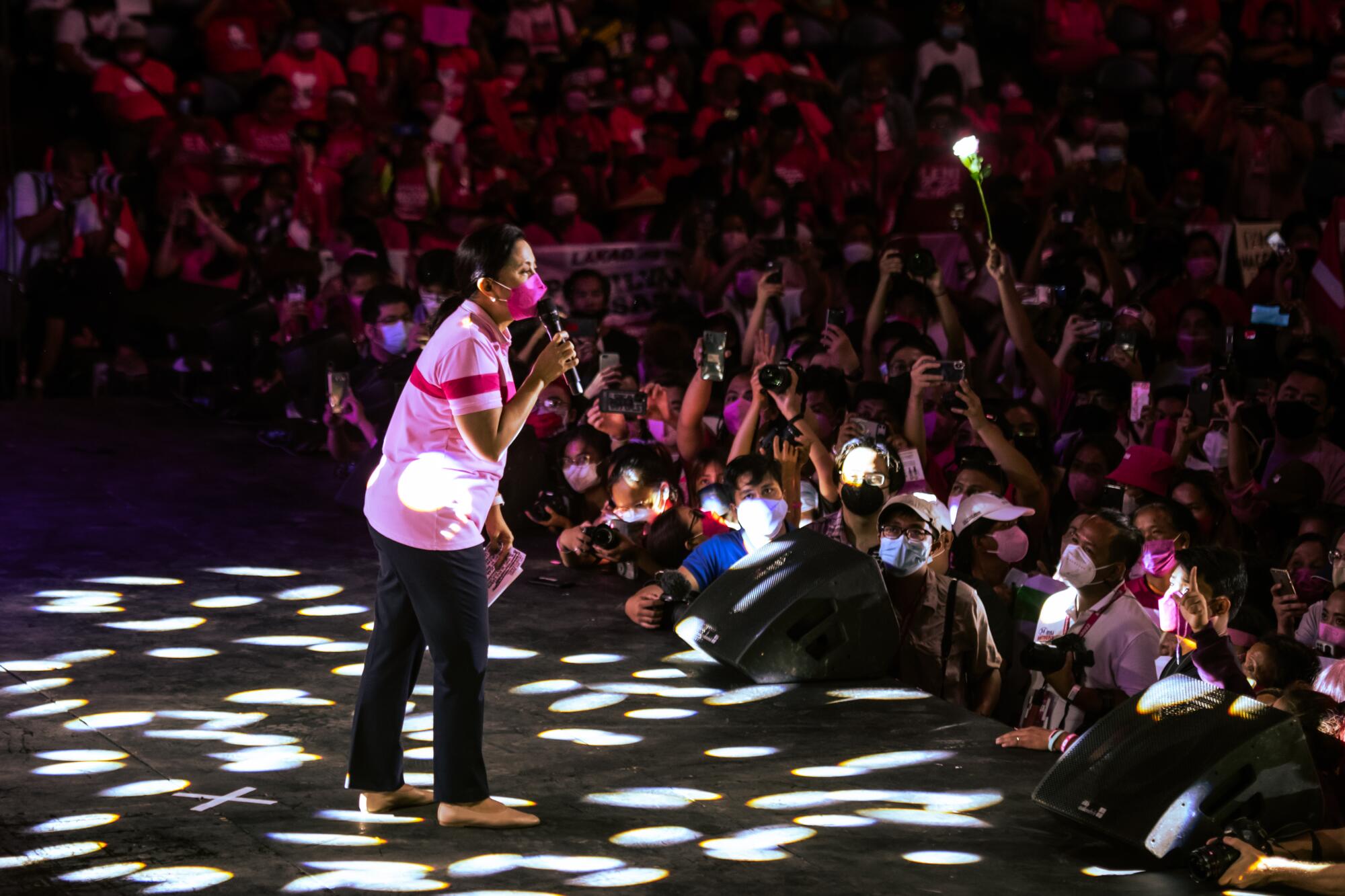 A woman in a pink mask and pink top holds a microphone as she stands before a crowd 