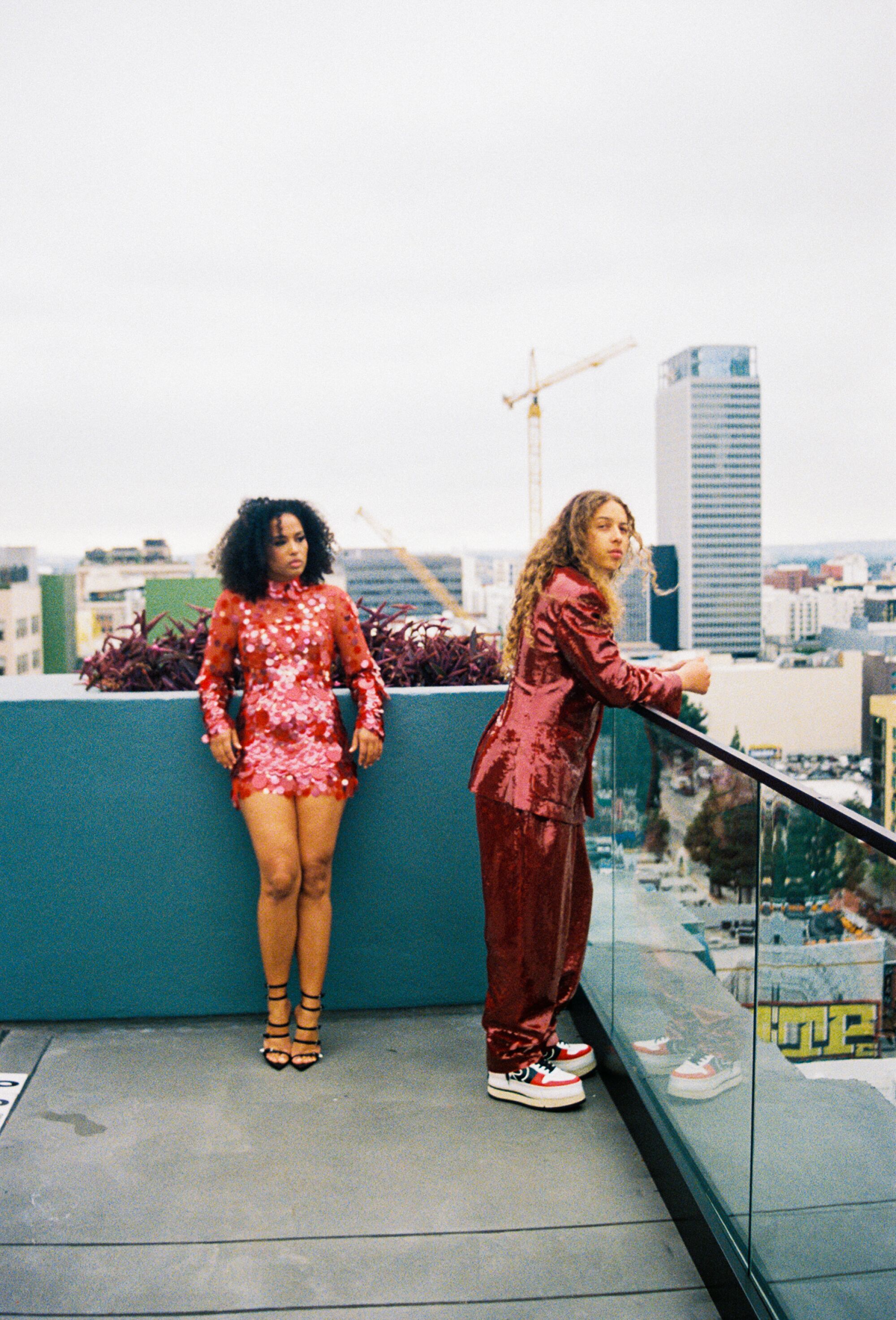 Two people wearing sequined red outfits lean against a waist-high wall on a rooftop.